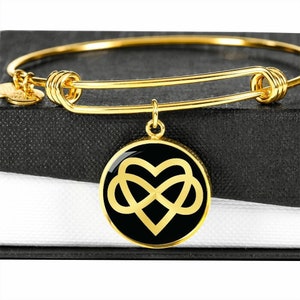 Puede incluir: Un brazalete de oro con un dije negro que presenta un símbolo de infinito dorado entrelazado con un corazón.