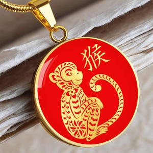 Może przedstawiać: Złoty naszyjnik z wisiorkiem z czerwoną, okrągłą zawieszką. Zawieszka przedstawia złoty wzór małpy i chińskie znaki. Małpa jest szczegółowo zdobiona kwiatowymi wzorami. Naszyjnik ma złoty łańcuszek.