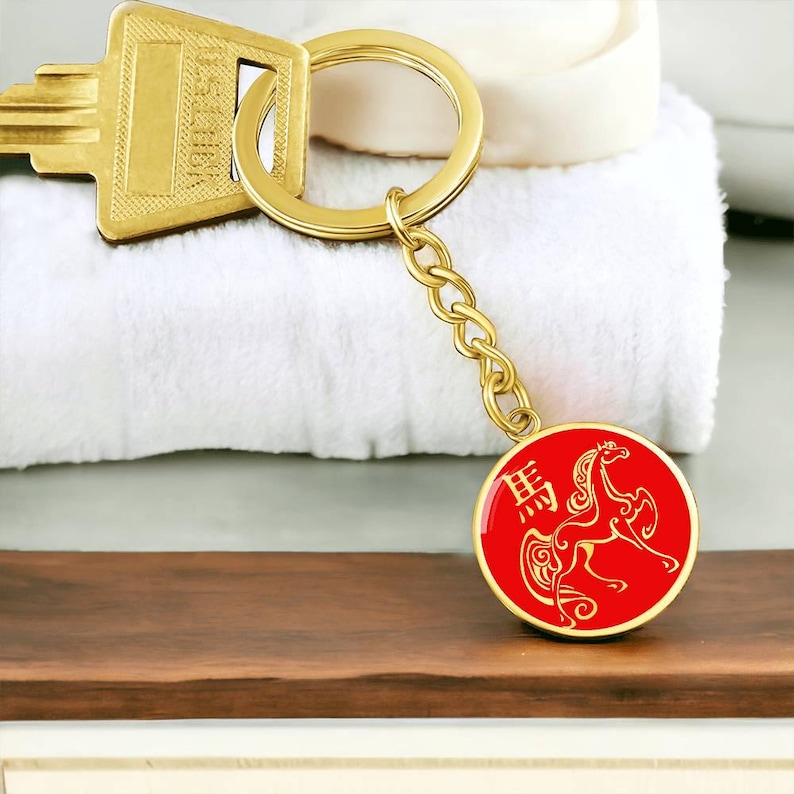 May include: A gold-coloured keyring with a key and a round charm. The charm is red with a gold horse design and Chinese characters. The keyring is on a wooden surface with a white towel in the background.