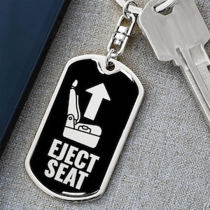 Puede incluir: Un llavero con placa de identificación negro y plateado con las palabras "EJECT SEAT" y un gráfico de flecha hacia arriba. El llavero está unido a una cadena plateada y una llave.