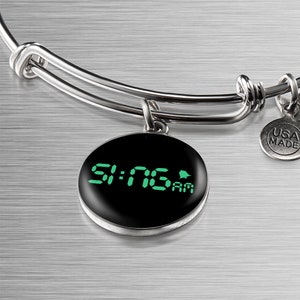 Op de afbeelding: Een zilveren armband met een ronde bedel met een zwarte achtergrond en een groene digitale klok die 9:16 AM laat zien. De bedel heeft ook een klein belpictogram. De armband heeft een "USA MADE"-tag.