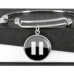 May include: A silver bangle bracelet with a round charm featuring a black and white pause symbol.