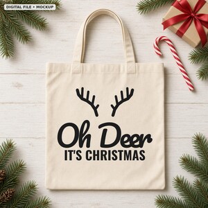K&ouml;nnte beinhalten: Eine beige Stofftasche aus Segeltuch mit einer schwarzen Grafik von Hirschgeweihen &uuml;ber dem Text Oh Deer IT'S CHRISTMAS. Die Tasche hat zwei Tragegriffe und ist auf einer hellen Holzoberfl&auml;che mit Tannenzweigen, einem Geschenk und einer Zuckerstange.