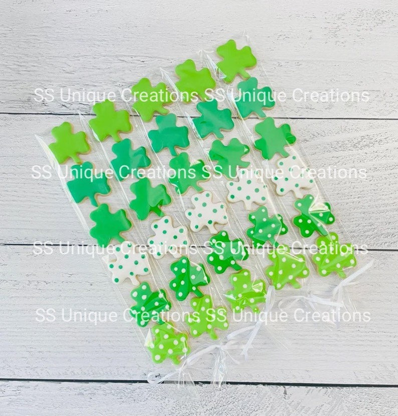 May include: Six clear cellophane bags filled with green and white shamrock-shaped cookies. The cookies are decorated with green and white polka dots. The bags are tied with white ribbon. The text "SS Unique Creations" is printed on the bags.