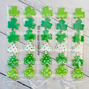 May include: Five clear cellophane bags filled with green shamrock cookies. Each bag contains five cookies, with the top cookie being solid green, the next two cookies being green with white polka dots, and the bottom two cookies being solid green. The bags are tied with white ribbon and the text "SS Unique Creations" is printed on the bags.