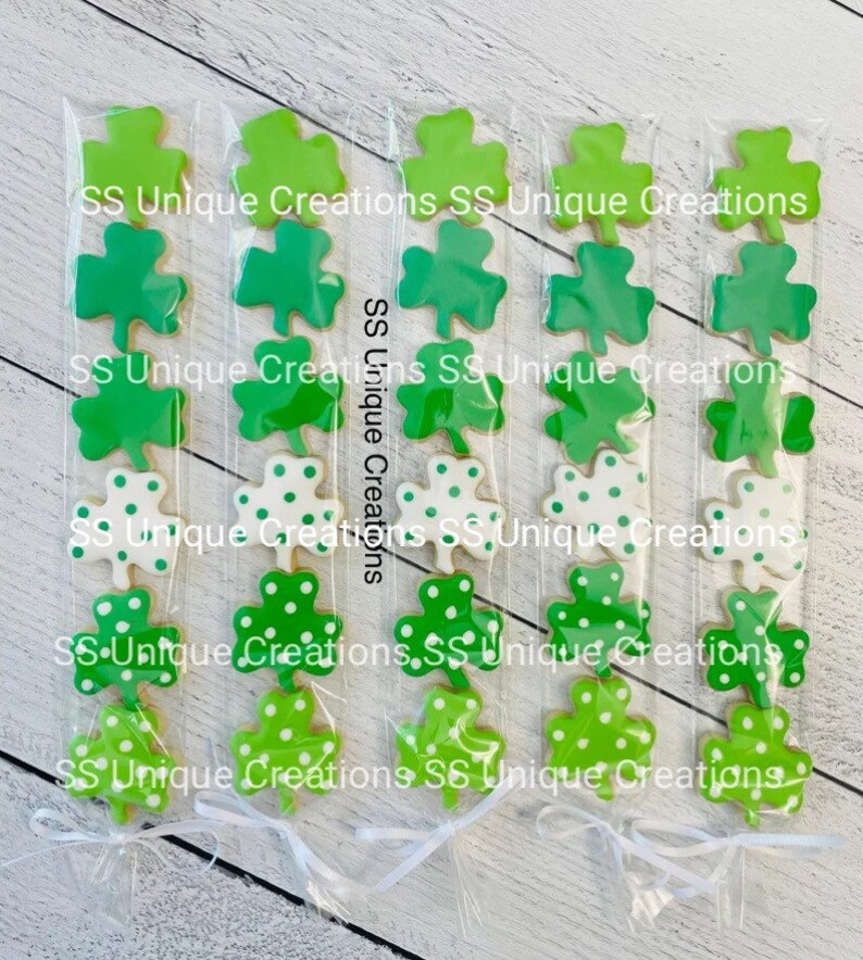 May include: Five clear cellophane bags filled with green shamrock cookies. Each bag contains five cookies, with the top cookie being solid green, the next two cookies being green with white polka dots, and the bottom two cookies being solid green. The bags are tied with white ribbon and the text "SS Unique Creations" is printed on the bags.