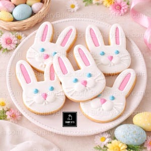 May include: White bunny-shaped sugar cookies with pink inner ears, blue eyes, and pink noses are arranged on a white plate. The cookies are decorated with white icing and sit on a table with Easter eggs and flowers.