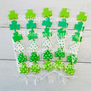 May include: Six clear cellophane bags filled with green shamrock-shaped cookies. The cookies are decorated with white polka dots. The bags are tied with white ribbon and the text "SS Unique Creations" is printed on the bags.