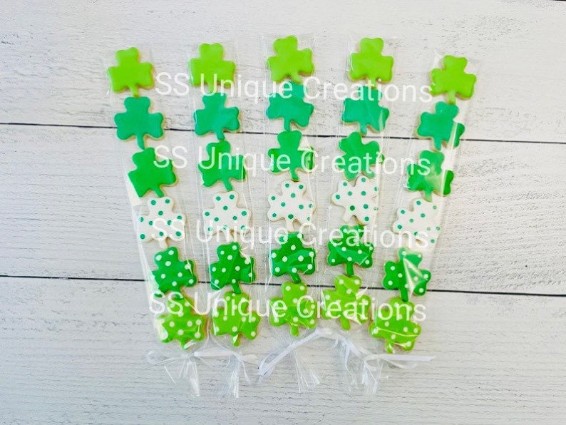 May include: Six clear cellophane bags filled with green shamrock-shaped cookies. The cookies are decorated with white polka dots. The bags are tied with white ribbon and the text "SS Unique Creations" is printed on the bags.