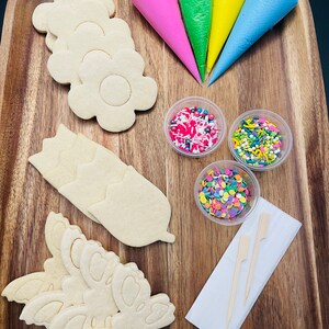 Op de afbeelding: Een houten dienblad met benodigdheden voor het versieren van koekjes. Er zijn vier spuitzakken gevuld met verschillende gekleurde glazuur, drie kommen met sprinkles, twee houten stokjes en zes suikerkoekjes in de vorm van bloemen en vlinders.