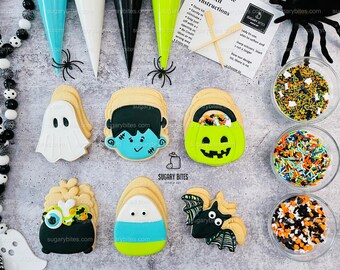 May include: A set of six Halloween-themed sugar cookies decorated with various characters, including a ghost, Frankenstein, a jack-o-lantern, a cauldron, a bat, and a candy corn. The cookies are arranged on a gray background with sprinkles in bowls.