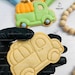 May include: A plain, unpainted sugar cookie in the shape of a pickup truck with a pumpkin in the bed. The cookie is being held by a gloved hand. The cookie is surrounded by other cookies and piping bags.