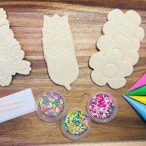 Op de afbeelding: Een koekjesdecoratieset met vier suikerkoekjes in de vorm van een vlinder, een bloem en een tulp. De set bevat vier spuitzakken met roze, groene, gele en blauwe frosting, drie bakjes met sprinkles en twee houten stokjes.
