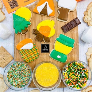 May include: A collection of camping-themed sugar cookies, including a tent, campfire, and s'mores, arranged on a wooden board. The cookies are decorated with colorful icing and sprinkles. Three small glass bowls contain yellow and multicolored sprinkles. The text "Sugary Bites" is visible.