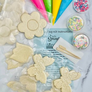 Op de afbeelding: Een lente-thema koekjesdecoratieset met verschillende koekjesvormen, waaronder bloemen en vlinders. De set bevat vier zakjes glazuur in roze, groen, geel en blauw, evenals drie bakjes met sprinkles. De instructies voor de set zijn zichtbaar op de afbeelding, met de tekst "hello spring".