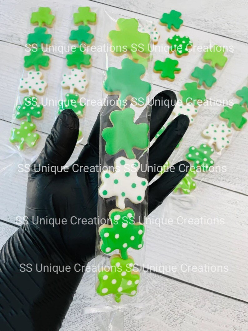 May include: A clear plastic bag filled with six green and white shamrock-shaped cookies. The cookies are decorated with green and white polka dots. The bag is held by a gloved hand.