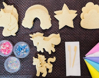 Op de afbeelding: Een assortiment suikerkoekjes in de vorm van eenhoorns, samen met regenboog-, ster- en wolkvormen. De afbeelding toont ook kleine bakjes met sprinkles, houten vorken en spuitzakken gevuld met kleurrijke glazuur.