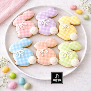 May include: A plate of Easter bunny-shaped cookies with pastel gingham patterns in pink, purple, yellow, blue, peach, and green. Each cookie has a white bow and a white pom-pom tail. Decorative Easter eggs surround the plate.