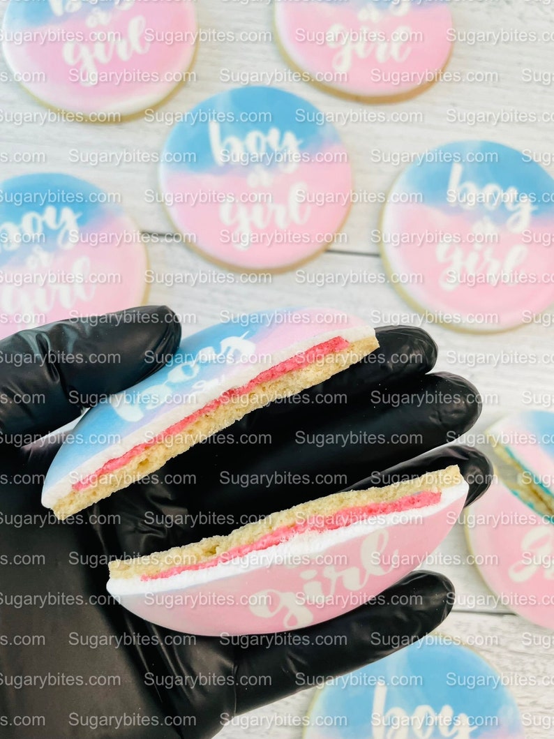 May include: Two sugar cookies decorated with blue and pink icing. The cookies are split in half to show the inside of the cookie. One cookie is decorated with the words "boy or girl" and the other cookie is decorated with the words "it's a girl".