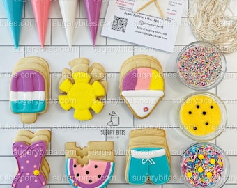 May include: A set of six decorated sugar cookies with summer themes, including a popsicle, a sun, a snow cone, a watermelon slice, a swimsuit, and swim trunks. The cookies are arranged on a white tiled surface with bowls of sprinkles and piping bags.
