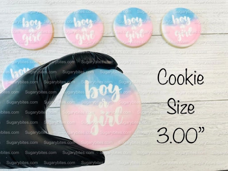 May include: Four decorated sugar cookies with blue and pink icing and the text "boy or girl" in white. A gloved hand holds a similar cookie in the centre of the image. The text "Cookie Size 3.00" is printed below the cookies.