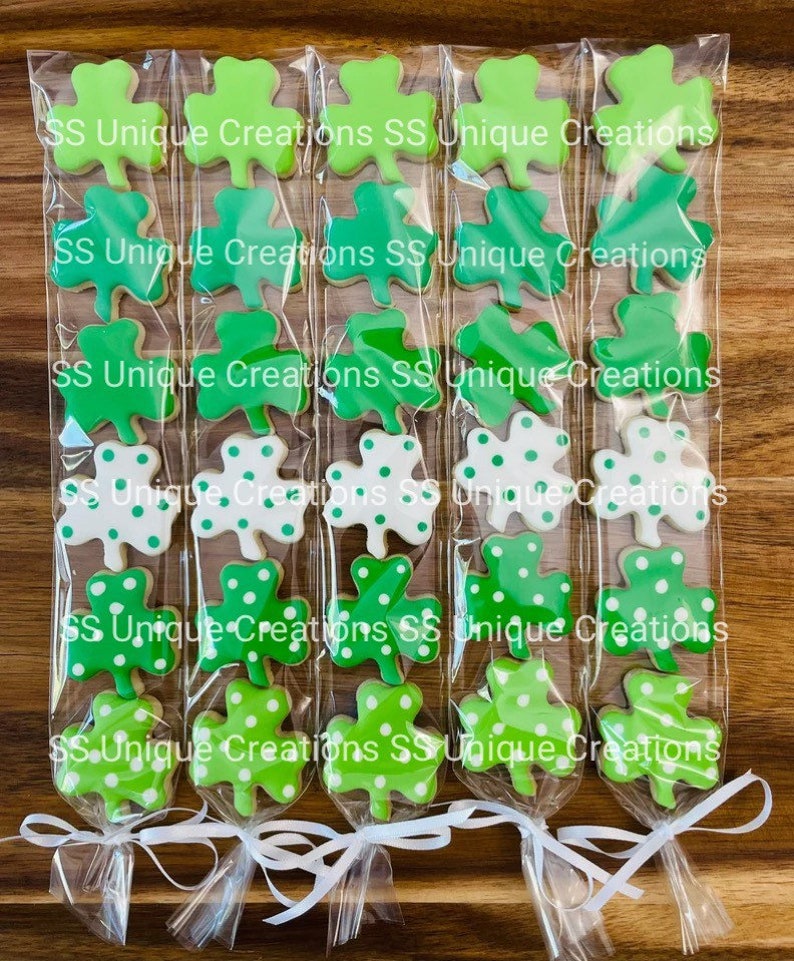 May include: Six clear cellophane bags filled with green and white shamrock-shaped cookies. Each bag is tied with a white ribbon and has the text "SS Unique Creations" printed on it.