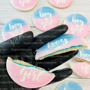 May include: A close-up of a hand holding several round sugar cookies decorated with blue and pink ombre frosting and the words "boy" and "girl" in white icing. The cookies are on a white wooden surface.