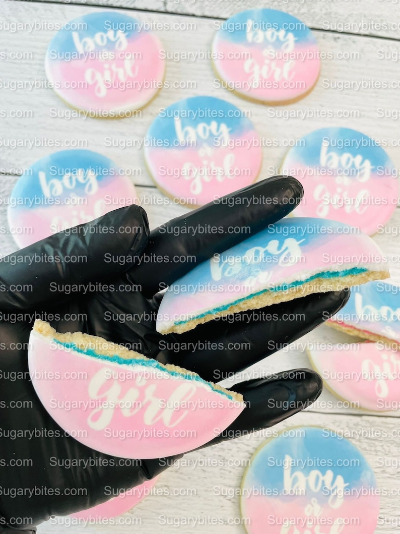 May include: A close-up of a hand holding several round sugar cookies decorated with blue and pink ombre frosting and the words "boy" and "girl" in white icing. The cookies are on a white wooden surface.