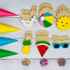 Può includere: Un set di stampini per biscotti colorati in varie forme, tra cui un ananas, un'anguria, occhiali da sole, un unicorno, un sole e una palla da spiaggia. Gli stampini per biscotti sono disposti su una superficie di legno bianco con sac à poche e codette.