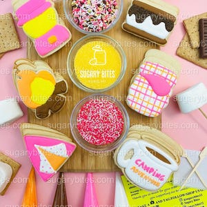 May include: Overhead shot of decorated sugar cookies and sprinkles on a wooden board. Cookies include a s'more, ice cream cone, and a mug with "Camping Adventure" text. Small bowls of sprinkles and icing cones are also visible.