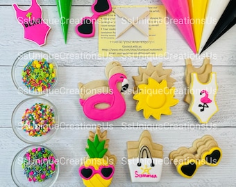 May include: A set of summer-themed cookie cutters, including a flamingo float, a sun, a swimsuit, a pineapple, a beach bag, and sunglasses. The cutters are made of white plastic and are arranged on a wooden surface. There are also three bowls of colorful sprinkles.