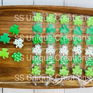 May include: Four clear bags filled with green and white shamrock-shaped cookies. The bags are tied with white ribbon and the text "SS Unique Creations" is printed on the wooden surface.