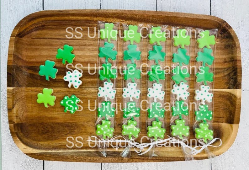 May include: Four clear bags filled with green and white shamrock-shaped cookies. The bags are tied with white ribbon and the text "SS Unique Creations" is printed on the wooden surface.