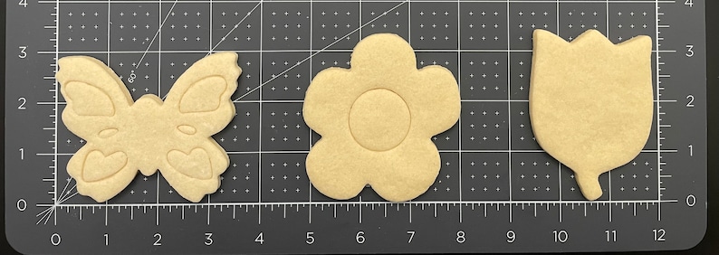 Op de afbeelding: Drie houten koekjesvormen in de vorm van een vlinder, een bloem en een tulp. De vormen zijn beige van kleur en zijn gerangschikt op een zwart-wit raster.