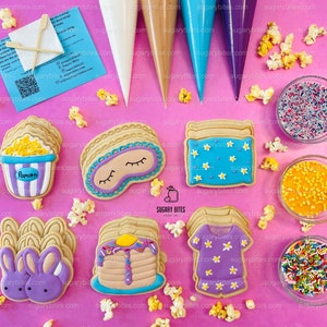 May include: A set of six decorated sugar cookies with various themes, including popcorn, a sleep mask, a pyjama top, a stack of pancakes, and two bunnies. The cookies are arranged on a pink surface with sprinkles, popcorn, and piping bags.
