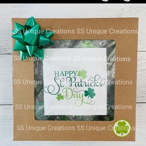 May include: A brown cardboard box with a clear window showing green and white candy. The box is decorated with a green bow and a green shamrock sticker. The text "Happy St. Patrick's Day" is visible in the window.