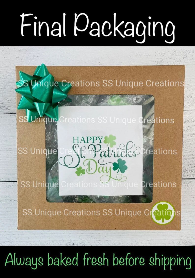 May include: A brown cardboard box with a clear window showing green and white candy. The box is decorated with a green bow and a green shamrock sticker. The text "Happy St. Patrick's Day" is visible in the window.