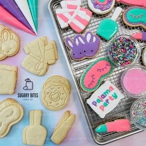 May include: A metal baking rack with various decorated sugar cookies, including a bunny, a gift box, a pillow, a sleep mask, a lipstick, and a nail polish bottle. The cookies are decorated with pink, purple, blue, and green icing. There are also bowls of sprinkles and sugar on the rack. The text "Pajama Party" is written on one of the cookies.