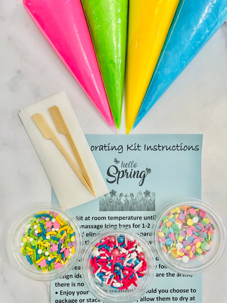 Op de afbeelding: Een lente-thema taartdecoratieset met vier spuitzakken in roze, groen, geel en blauw, twee houten decoratiestockjes en drie bakjes met kleurrijke sprinkles. De instructies zeggen "Hello Spring" en bevatten instructies voor het gebruik van de set.