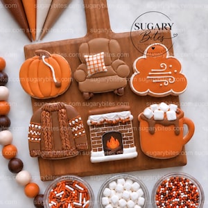 Könnte beinhalten: Sechs verzierte Kekse auf einem Holzbrett: ein orangefarbener Kürbis, ein brauner Sessel mit kariertem Kissen, eine orangefarbene Wolke mit Windwirbeln, ein brauner Strickpullover, ein Kamin aus Ziegelsteinen und ein Becher Kakao mit Marshmallows.