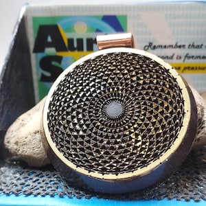 Pode incluir: Um pingente redondo dourado e preto com um design geométrico. O pingente está em uma corrente de cobre.