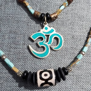 May include: A turquoise and silver Om pendant necklace with a black and white patterned bead. The necklace is made of brown, beige, and turquoise beads.
