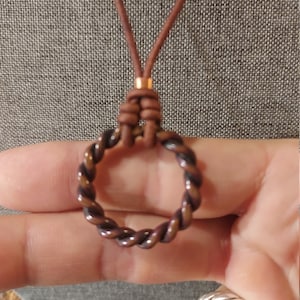 Puede incluir: Un collar con un colgante de alambre de cobre trenzado en forma de círculo y un cordón de cuero marrón.