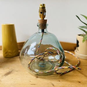 May include: A clear, rounded glass bottle lamp base with a cork stopper and a brass light socket. A multi-colored, braided fabric electrical cord is coiled inside the transparent glass body and extends out from the top of the bottle.