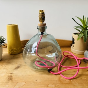 May include: A clear glass lamp base with a rounded, bulbous shape. It features a brass-colored metal light socket at the top, secured with a cork stopper. A bright pink electrical cord is coiled inside the glass base and extends outward.