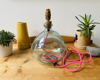 May include: A clear glass lamp base with a rounded, bulbous shape. It features a brass-coloured metal light socket at the top, secured with a cork stopper. A bright pink electrical cord is coiled inside the glass base and extends outward.