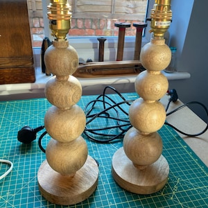 May include: A pair of table lamp bases crafted from light-colored wood, each featuring a vertical stack of four spherical shapes mounted on a circular wooden base. Each lamp includes a metallic gold-toned socket at the top with a black power cord.
