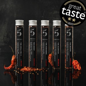Pode incluir: Cinco tubos de vidro contendo diferentes tipos de flocos de pimenta. Cada tubo tem um rótulo preto com o nome da pimenta, a escala de Scoville e as notas de degustação. Os tubos estão dispostos em uma fileira sobre uma superfície preta. Os flocos de pimenta são vermelhos e marrons.
