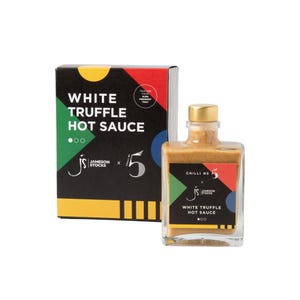 Può includere: Una bottiglia quadrata di salsa piccante al tartufo bianco con tappo dorato, accanto a una scatola nera con la stessa etichetta. L'etichetta include il testo "White Truffle Hot Sauce" e "Chilli No 5". La scatola ha un design geometrico colorato.