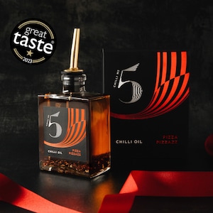 Puede incluir: Una botella cuadrada de vidrio de aceite de chile con un pico dorado y una etiqueta negra con el número 5 y las palabras "Chilli Oil" y "Pizza Pizzazz". Una caja negra con el mismo diseño está detrás de la botella. Una pegatina "Great Taste 2023" está en la parte superior izquierda.